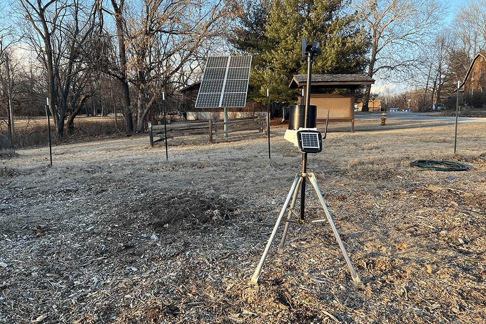 A solar-powered tripod stands in a field to collect weather data, with a large solar panel in the background also used to power the weather collectors. 